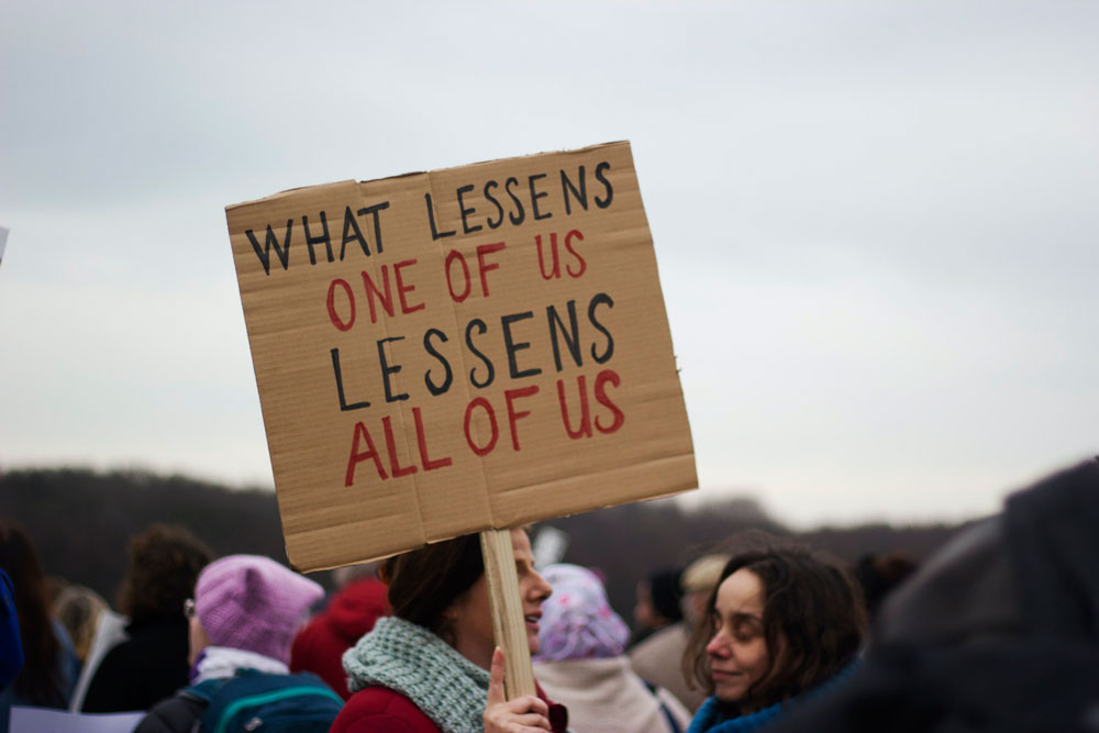 women protesting with sign "what lessens one of us lessens all of us"
